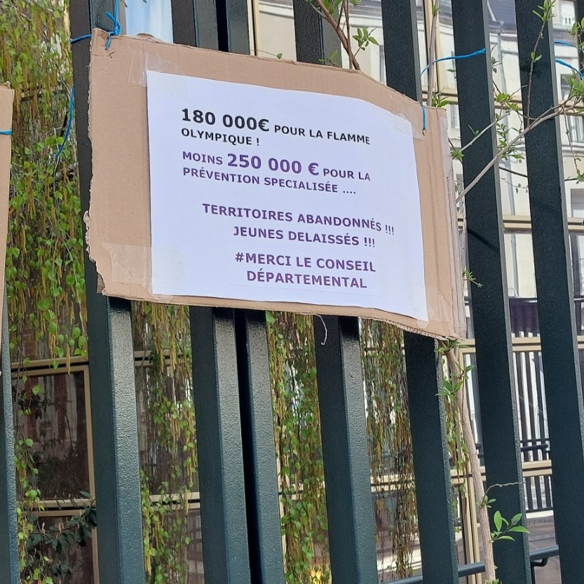 Pancarte de protestation accrochée aux grilles de l'hôtel du département : 180 000 € pour la flamme olympique ! =Moins 250 000 € pour la prévention spécialisée. Territoires abandonnés !!! Jeunes délaissés !!! #Merci le Conseil départemental